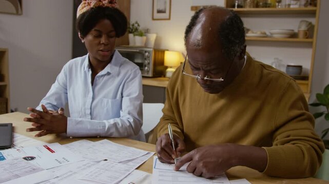 Medium shot of Black senior man writing checks and paying bills while sitting at table with documents scattered, young woman helping