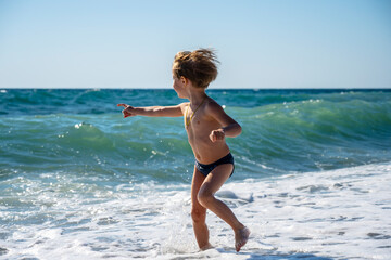 Fototapeta premium Young child runs along sunny beachfront with excitement, Energetic young explorer sports swim trunks while leaping near foamy ocean waves