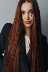 Woman with long straight brown hair wearing black blazer and white shirt posing against gray...