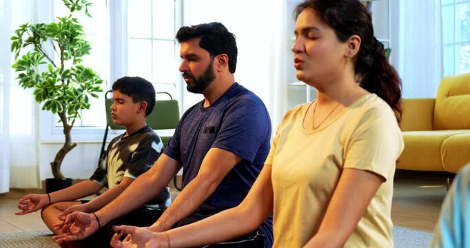 Indian young family doing omkar meditation pranayama at home chanting aum breathing yoga together indoors during peaceful morning hours in a modern living room promoting yogic lifestyle