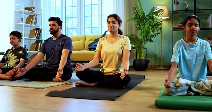 Indian young family doing omkar meditation pranayama at home chanting aum breathing yoga together indoors during peaceful morning hours in a modern living room promoting yogic lifestyle