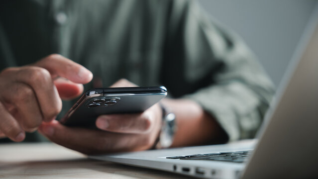 Close-up of a person's hand holding a smartphone with a laptop in the background. The image focuses on the smartphone's camera lens.