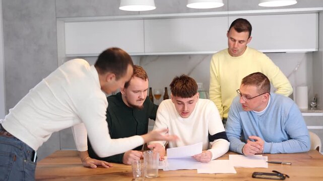 Upset worried man sitting at table in home kitchen with papers in hands, reading unexpected medical results with group of male friends solacing and comforting him 
