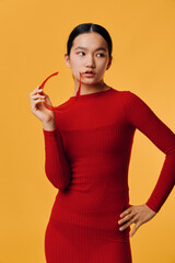Young woman in red dress holding red glasses near face looking to side posing on orange background....