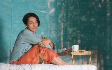 Mindfulness and feel-good moments. middle aged woman rests while sitting on a bed near a window in a country house in the forest. solo retreat