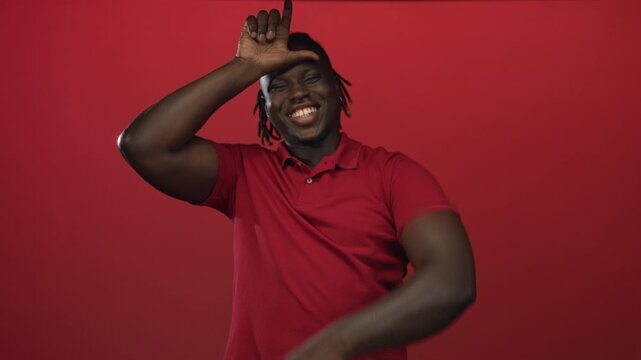 Man with hand on forehead forming l gesture and pointing finger toward camera while smiling in a red studio backdrop and wearing a red polo shirt; playful confidence.