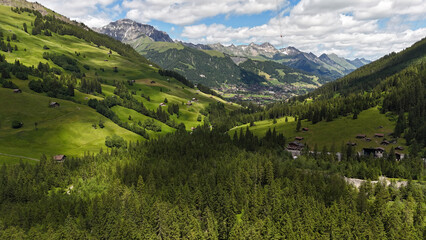 Obraz premium Drone view, aerial of hiking path into beautiful valley, pine tree forest in foreground. Adelboden village in background. Swiss mountains, beautiful alpine nature, landscape. Tranquility, pure pine tr