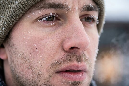A man stands in a cold area with frost on his face. He looks serious while the snow surrounds him in a winter scene. His breath is visible in the chilly air