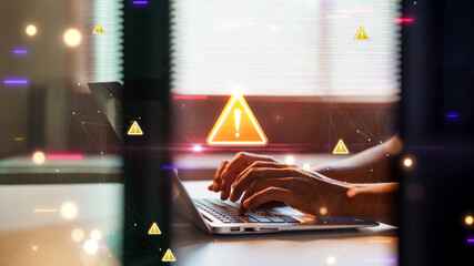 close up of hands typing on a laptop keyboard at a desk, featuring a glowing digital warning sign overlay representing cybersecurity threats, system errors, or data privacy alerts.