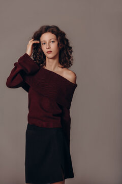 The model is a young beautiful brunette girl with curls on a gray background in a black short skirt and a bard sweater