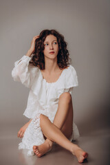 Fototapeta premium The model is a young beautiful brunette girl with curls on a gray background in a white blouse and lace skirt