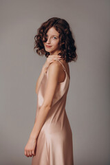 Fototapeta premium The model is a young beautiful brunette girl with curls on a gray background in a pink silk dress