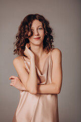 Fototapeta premium The model is a young beautiful brunette girl with curls on a gray background in a pink silk dress