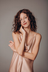 Fototapeta premium The model is a young beautiful brunette girl with curls on a gray background in a pink silk dress
