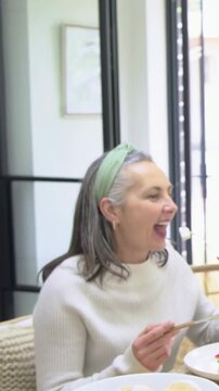 Vertical video: Chatting senior woman in white sweater reaching for bowl at wooden table, eating