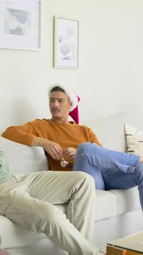 Vertical video: Smiling man in Santa hat fidgeting ornament on sofa while companion entering