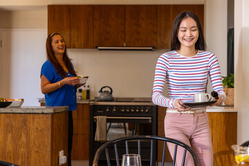 Asian mother and daughter carrying tea tray and holding food bowl by kitchen island