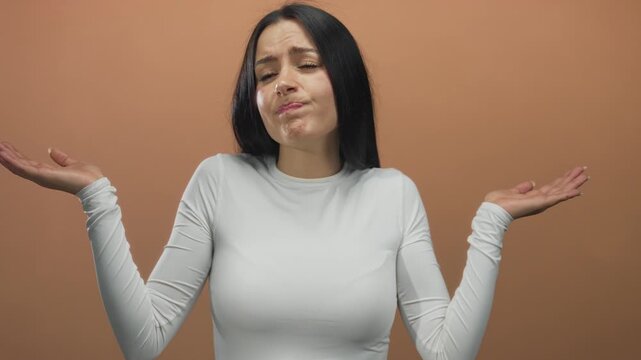 Woman in white shirt with puzzled expression against brown background shows confusion and uncertainty with her hands raised in questioning gesture.