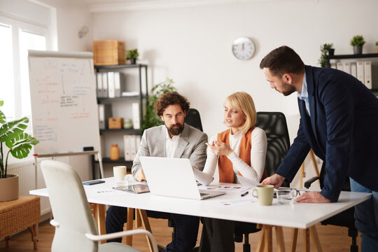 A group of three people discusses strategies while sitting at a table with a laptop in a well-lit office space. They focus on teamwork and planning for their project.