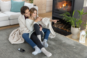 Naklejka premium Couple snuggling together in cream blanket on gray rug in front of fireplace holding white mug