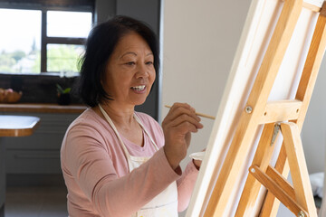 Senior asian woman painting blank canvas on wooden easel in art nook with paintbrush, palette