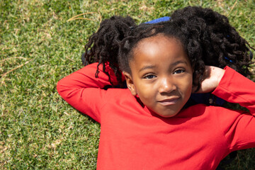 African american school-age girl lying on green lawn in red long-sleeve shirt under sunny sky