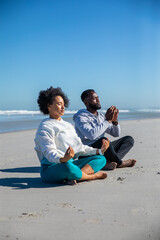 African american couple meditating cross-legged at water's edge on ocean beach in activewear