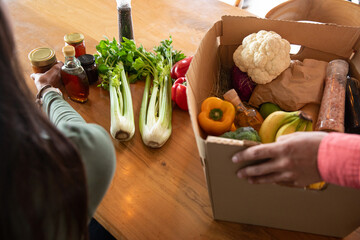 Diverse couple grasping cardboard box of produce, reaching jars at kitchen table, copy space © wavebreak3