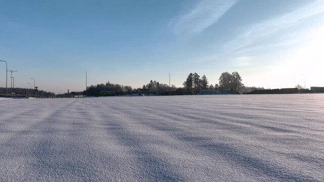 A vast snowy field is illuminated by the soft light of sunrise, trees and distant buildings visible on the horizon. Smooth panning camera movement