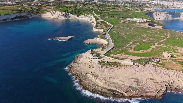 Forward-moving aerial drone flight along the southern coast of Malta, revealing rocky shoreline, coastal salt pans, winding roads, and expansive Mediterranean seascape near Triq Xrobb L-Għaġin.