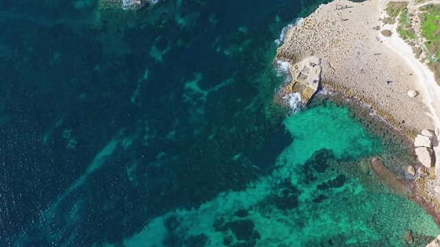 Top-down drone view of It-Taqtiegħa and the nearby rocky shoreline at Triq Xrobb L-Għaġin, highlighting turquoise Mediterranean waters, rugged limestone formations and Malta&rsquo;s pristine southern coast.