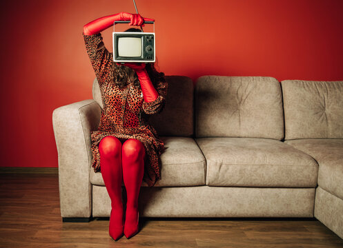 Woman in leopard dress with TV on head sitting on sofa in retro living room