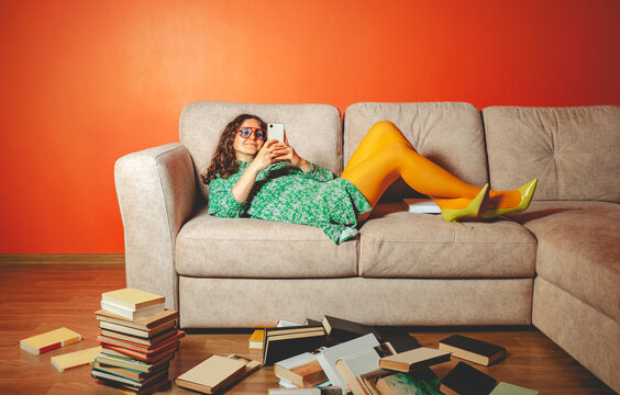 Person lying on sofa distracted by smartphone surrounded by books