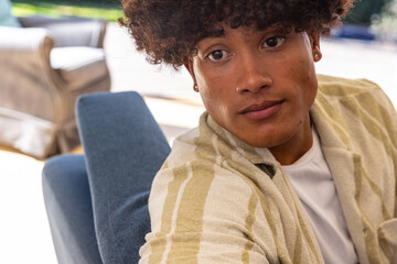Man wearing stud earrings sitting on lounge chair beside beige sofa in sunny patio light