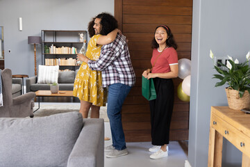 Diverse friends hugging and exchanging green gift bag in modern living room, pastel balloons