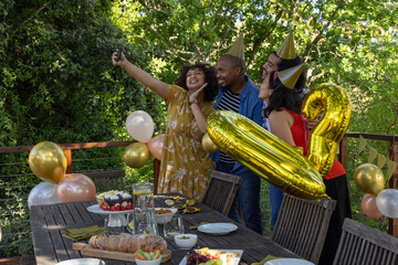 Obraz premium Diverse friends leaning wearing party hats posing with smartphone, balloons on wooden deck
