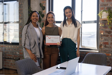 Obraz premium Diverse female coworkers holding framed certificate and smiling in loft office room with laptop