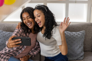 Obraz premium Diverse mom and teenage daughter smiling while holding smartphone and posing on couch at home