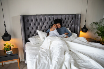 Diverse couple lying under white duvet on bed with tufted headboard, pendant lamps