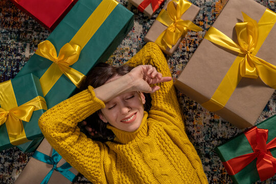 Smiling woman in yellow sweater lying on floor with holiday gifts indoors