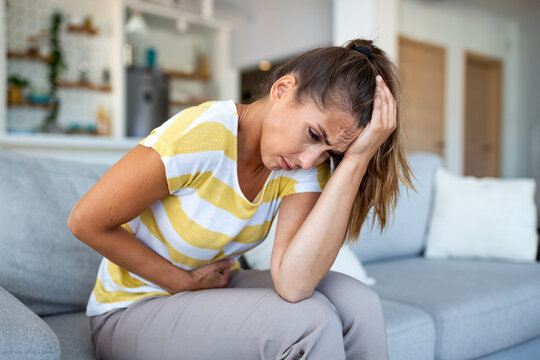 Woman holding head and stomach in pain sitting on couch. Migraine, abdominal cramps and PMS symptoms concept. Young female suffering from severe discomfort and illness at home. Healthcare.
