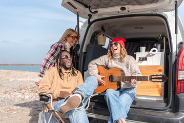 Diverse multiethnic Friends laughing and playing guitar during road trip at beach with camper van