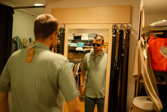 Young man trying on clothes in a fitting room and looking at his reflection in a mirror. Shopping, fashion choice, lifestyle and menswear concept.
