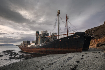 Fototapeta premium Vintage whaling vessels beached on the Icelandic shoreline. Weathered industrial ships abandoned near the ocean under a dramatic sky.