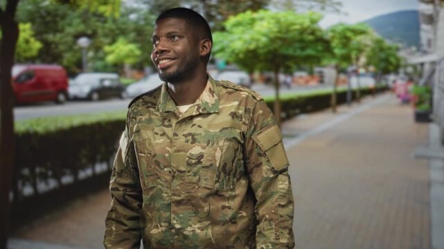 Soldier man smiling and looking up on a street in camouflage uniform and jacket, standing on a sidewalk near trees and parked cars; pride duty.