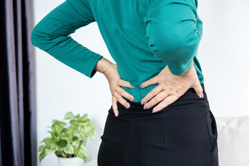 Middle-aged woman suffering from lower back pain or kidney ache at home. Mature female with hands on waist feeling spinal injury, muscle strain, or office syndrome discomfort