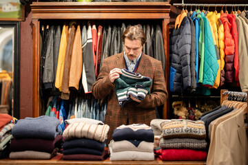Obraz premium Chic dressed man customer in vintage boutique choosing knitted jumper from display, mustachioed male buyer holding desired apparel item and checking examinees quality of clothing, deciding on trying.