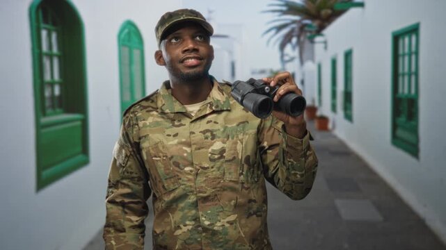 Soldier man holding binoculars on a whitewashed street with green shutters and a palm tree in view; duty watching vigilance.