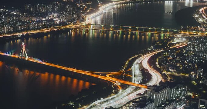 Aerial perspective captures illuminated bridges and busy traffic flowing along roads at night with reflections on the water surface. Time-lapse, Seoul, South Korea