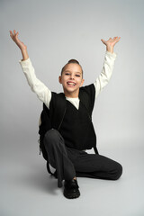 Joyful young girl in a school uniform and backpack crouching while laughing and throwing her hands up in celebration. Studio portrait on a neutral grey background